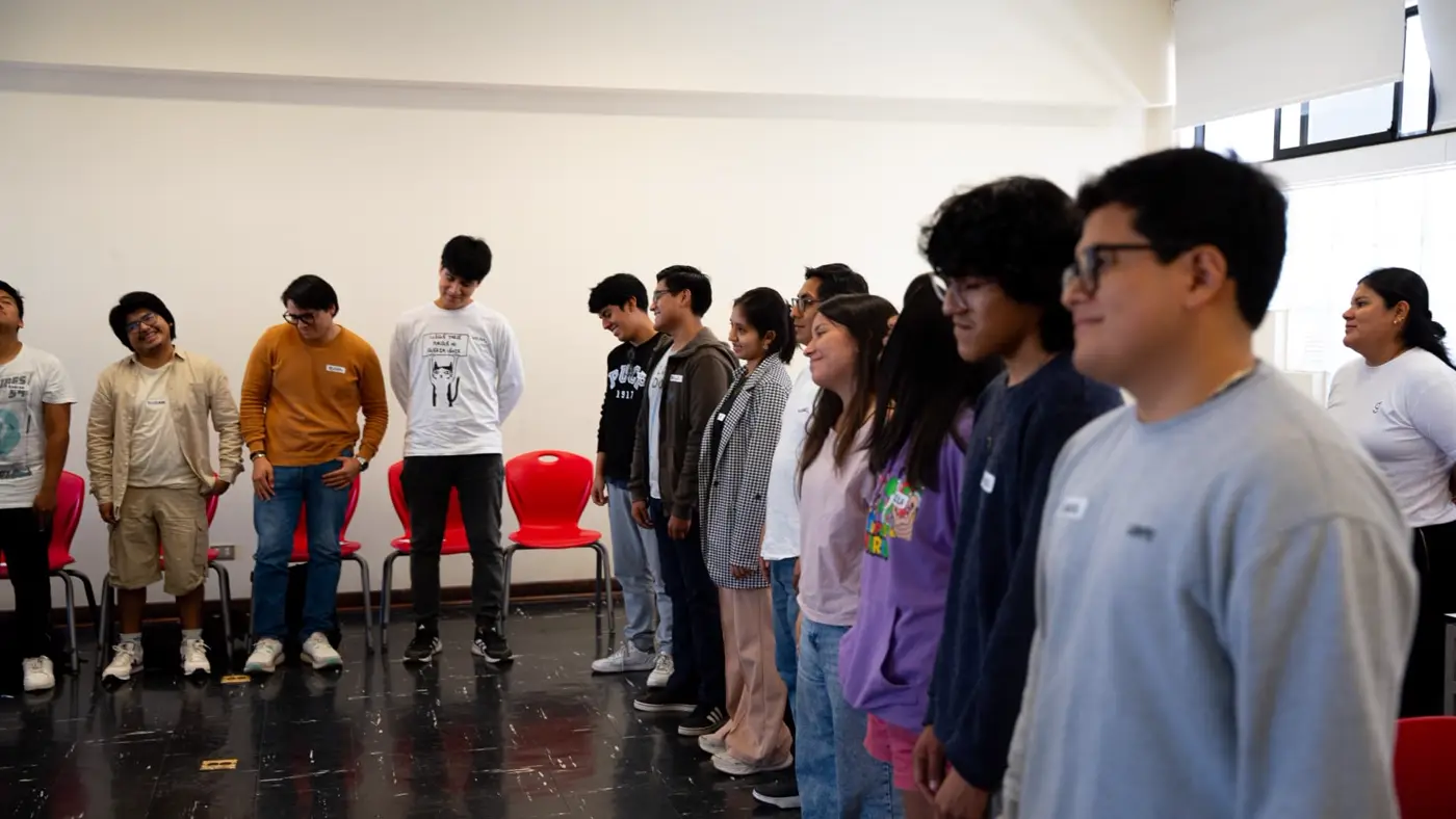 Estudiantes de pie en fila frente a sillas rojas durante una dinámica